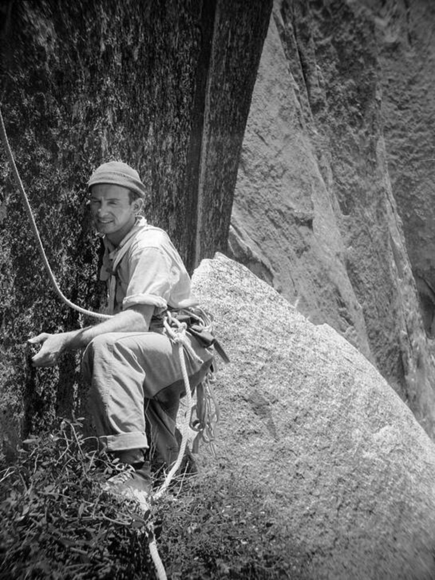 Black and white photo of a person climbing on a rock face, with a helmet and harness, in a mountainous wilderness setting.