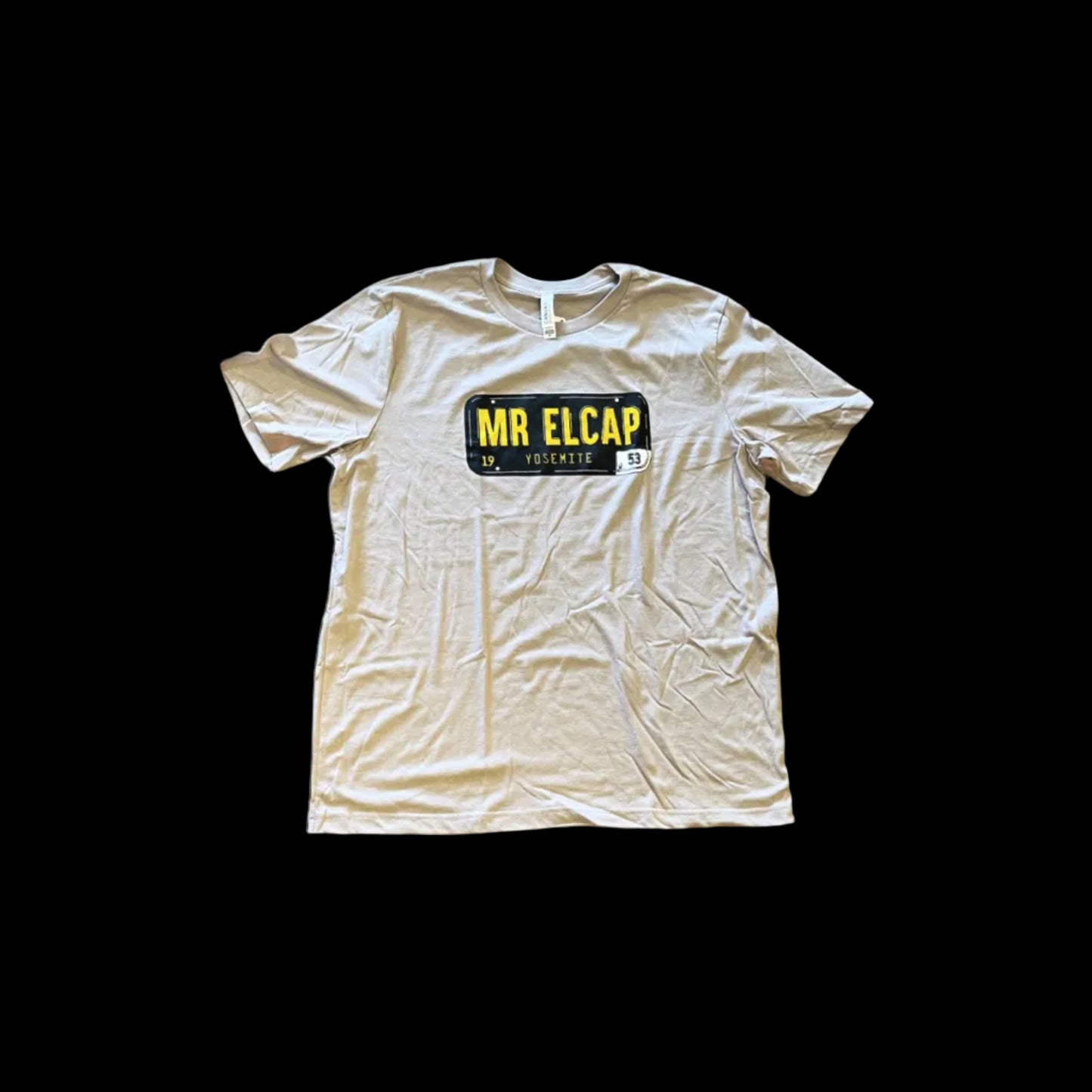 A beige t-shirt with 'MR ELCAP' printed in black text on the front, displayed on a wooden surface.